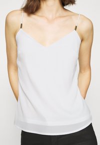 Femme portant un haut blanc sans manches à col en V avec de fines bretelles et des accents métalliques, associé à un pantalon noir, debout les mains dans les poches.