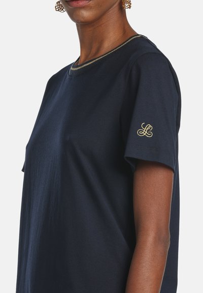 Nő zuhanszkék, rövid ujjú ingben, arany hímzett "L" betűvel az ujjon, és arany szegélyű nyakkivágással, vállától a felső mellkasig látható.