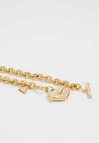 Gouden kettingarmband met een hartletterhanger met rhinestone accenten. Inclusief een driehoekig label met de merknaam en een toggle-sluiting.