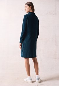 Abito in corda teal con maniche lunghe e colletto alto, lungo fino al ginocchio con una sottile texture a coste. Indossato con sneakers bianche e calzini.