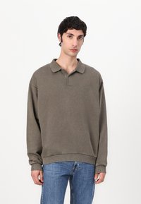 Oversized, long-sleeve polo in muted olive green cotton, featuring a buttonless collar and ribbed cuffs. Light texture with subtle branding.