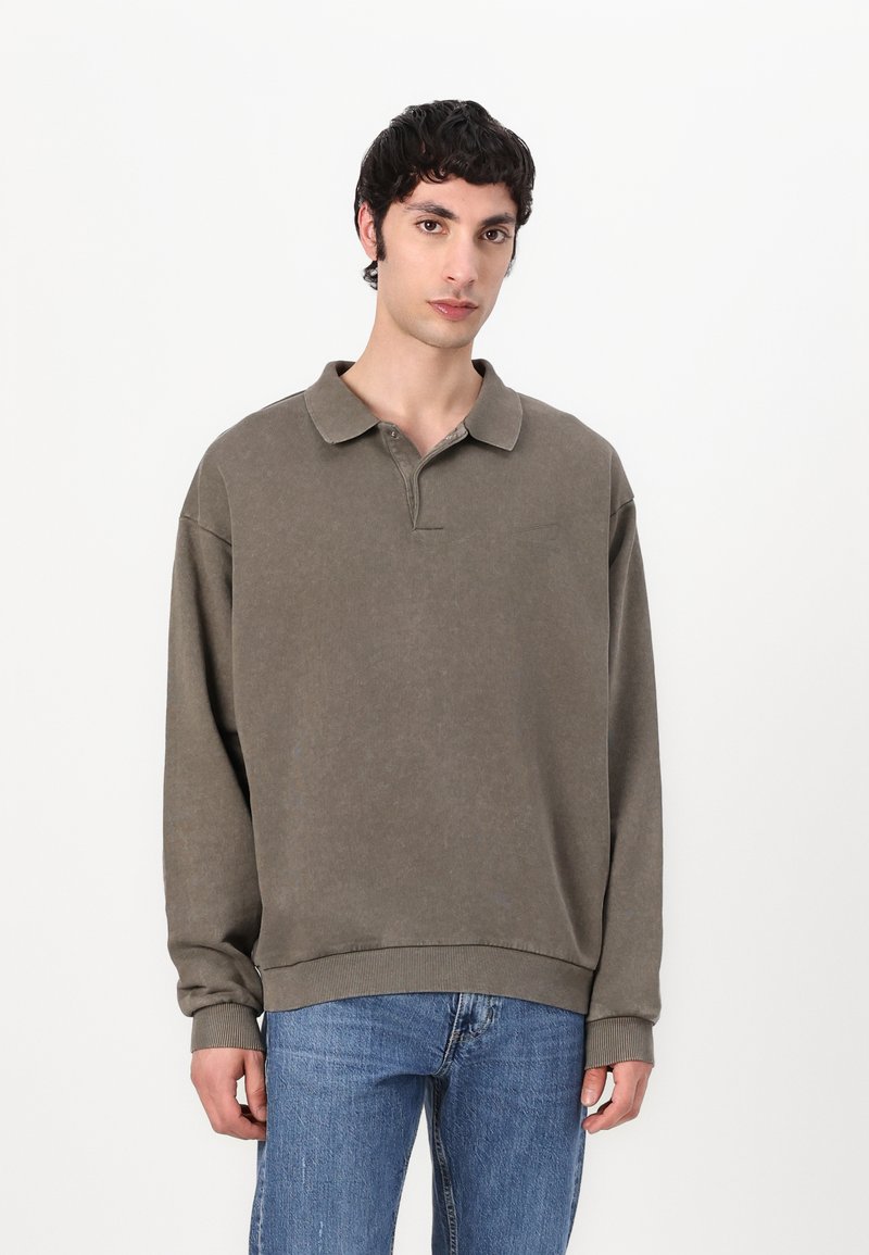 Oversized, long-sleeve polo in muted olive green cotton, featuring a buttonless collar and ribbed cuffs. Light texture with subtle branding.