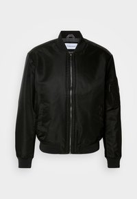 Blouson aviateur noir en matière brillante et lisse. Il présente une fermeture zippée, des poignets et un ourlet côtelés, ainsi qu'une poche sur la manche gauche.