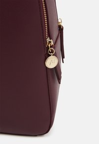 Gros plan sur le coin d'un sac en cuir bordeaux avec une fermeture éclair dorée et un pendentif rond en or gravé des initiales stylisées "A7" sur la languette de tirette.