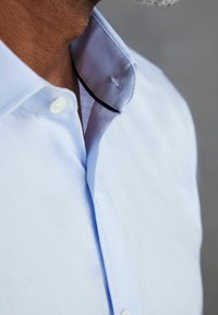 Hellblaues Hemd mit Knopfleiste und texturierter Webung, das einen kontrastierenden, sanften blauen Kragen sowie einen schwarzen Akzentstich an der Kragennaht aufweist.