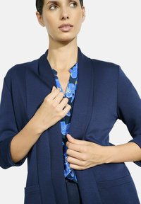 Tyrkysově modrá kimono stylová bunda vyrobená z texturované tkaniny, s třičtvrtečními rukávy a otevřeným předním částem, vrstvená přes modrou košili s květinovým potiskem.
