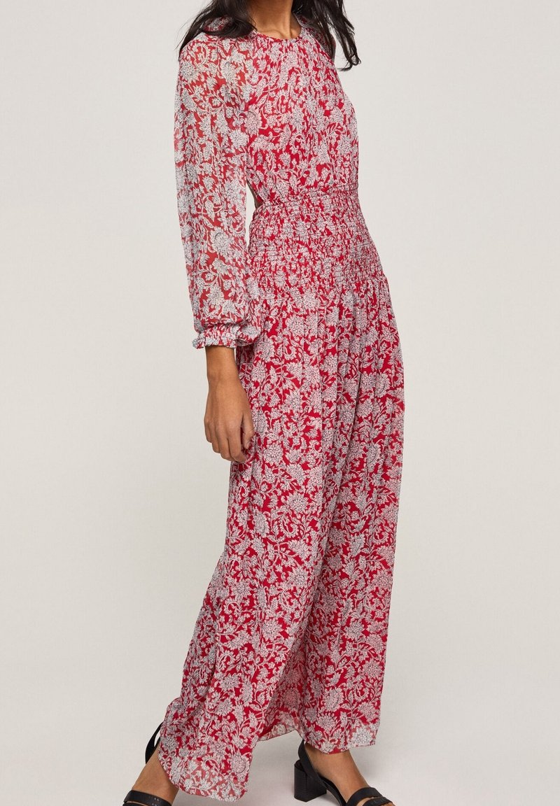 Femme portant une robe maxi rouge avec de longues manches transparentes et un motif floral blanc, associée à des sandales noires à talons blocs.