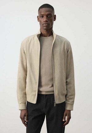 Hombre con chaqueta de ante beige con cremallera sobre un suéter marrón claro y pantalones negros, de pie frente a un fondo liso claro.