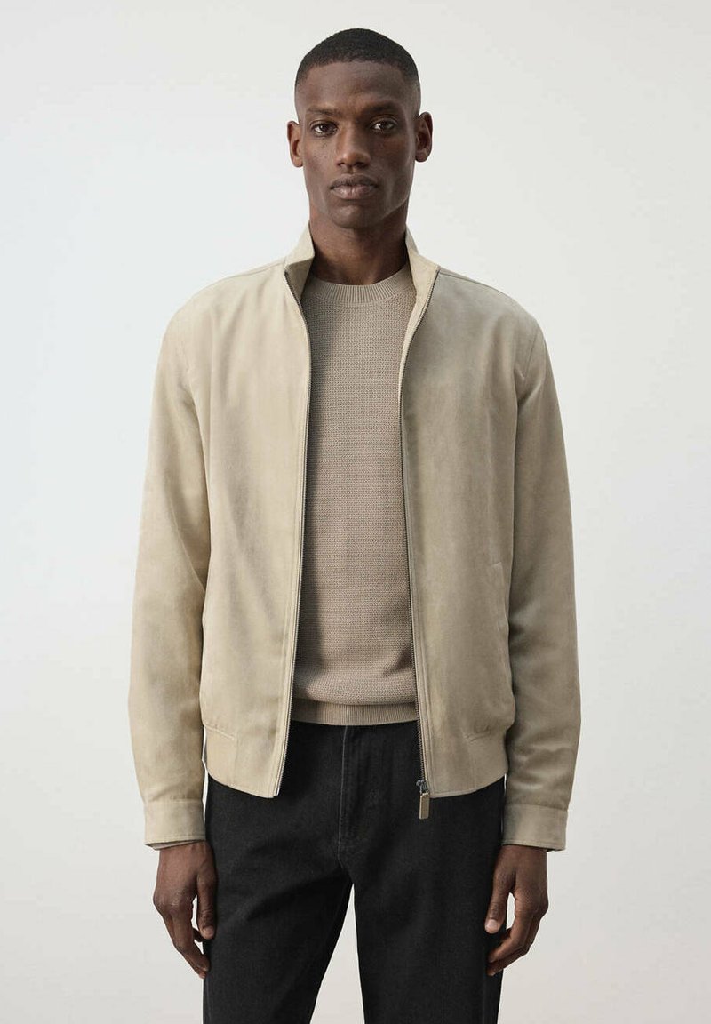 Homme portant une veste zippée en daim beige sur un pull marron clair et un pantalon noir, debout devant un fond clair uni.