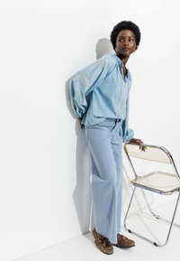 Chemise en denim bleu clair avec des motifs brodés, associée à un pantalon large bleu clair et des chaussures à imprimé léopard. Chaise transparente à proximité.