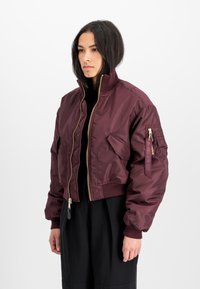 Kuttet maroon bomberjakke laget av skinnende nylon, med gullglidelås, ribbestrikkede mansjetter og en glidelåslomme på venstre erme.