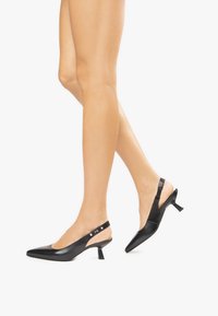 Tacchi slingback in pelle nera con punta affusolata, tacco quadrato basso e cinturino con dettagli metallici. Texture liscia, design minimalista.