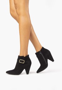 Černé suede kotníkové boty s špičatými špičkami, zúženými podpatky, zlatým přezkovým detailem a bočním zipem; texturovaný materiál s elegantním designem.