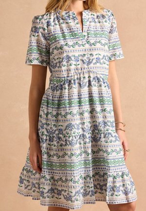 Femme portant une robe blanche à manches courtes avec des motifs floraux et rayés bleus et verts et un col en V, debout sur un fond beige.