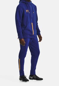 Felpa blu con dettagli arancioni, chiusura con zip e tasca a marsupio; pantaloni blu abbinati con striscia arancione laterale; indossati con scarpe da calcetto nere.