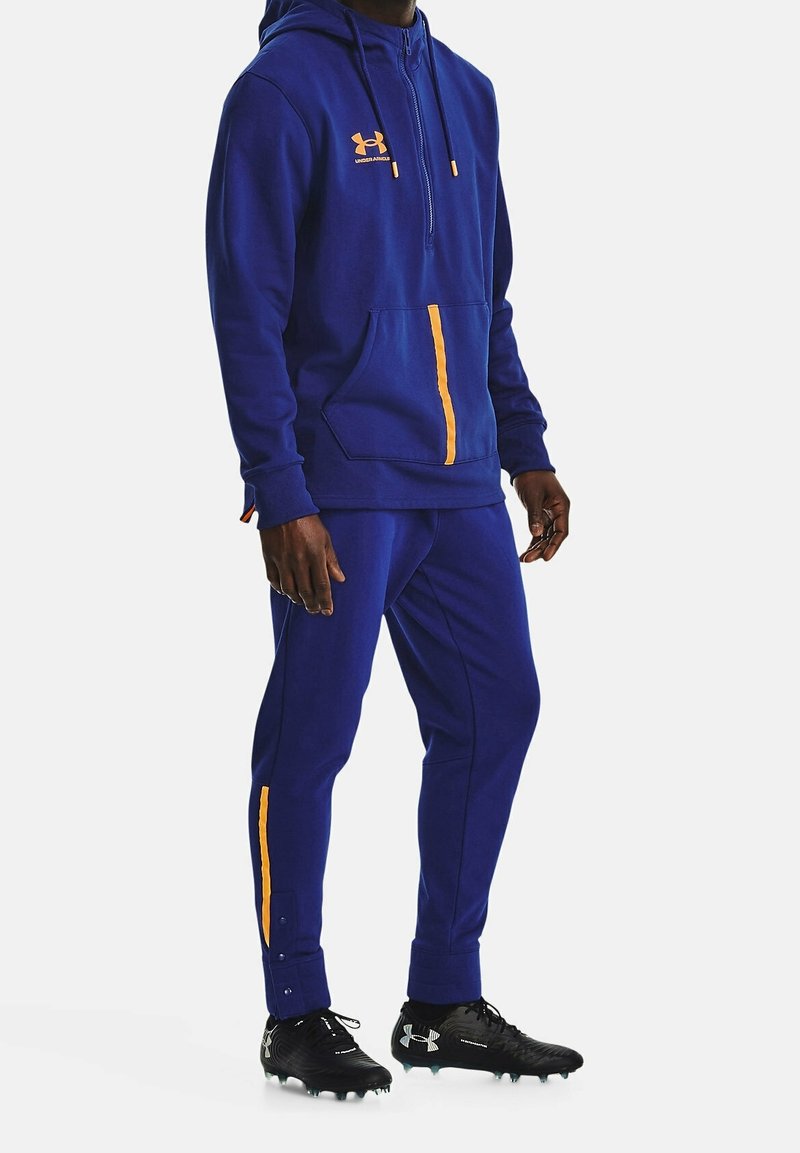 Felpa blu con dettagli arancioni, chiusura con zip e tasca a marsupio; pantaloni blu abbinati con striscia arancione laterale; indossati con scarpe da calcetto nere.