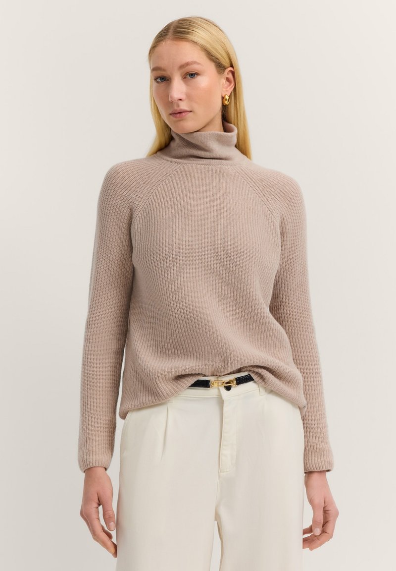 Beige gerippter Rollkragenpullover mit langen Ärmeln, der ein figurnahes Design und eine weiche Textur zeigt, kombiniert mit cremefarbenen Hosen und einem dunklen Gürtel.