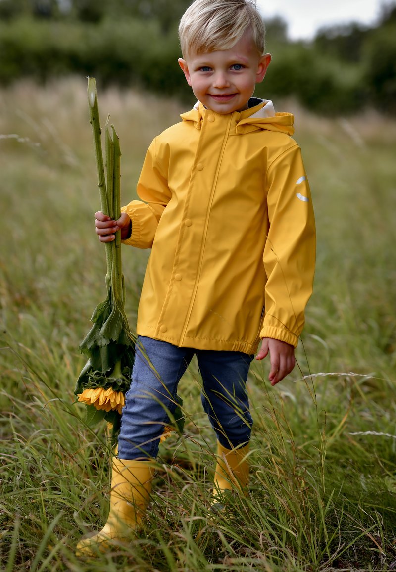 Sárga vízálló esőkabát kapucnival, szürke farmer és sárga gumicsizma. Napraforgókat tart a kezében, magas fűben áll.