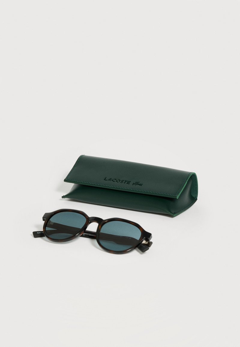 Sonnenbrille mit Schildpattfassung und dunklen Gläsern neben einem dunkelgrünen Lacoste-Schutzetui auf einer weißen Oberfläche.