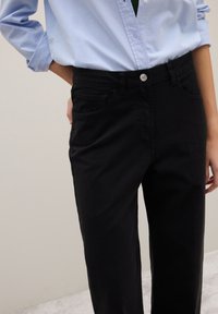 Pantaloni di cotone neri con due tasche frontali e una cintura con un solo bottone. Abbinati a una camicia celeste con bottoni, mostrando un look casual.