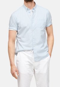 Světle modrá kostkovaná košile s krátkým rukávem, límečkem na knoflíky, přední kapsou a měkkou látkou, ve spojení s bílými lněnými kalhotami.