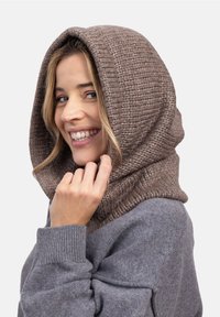 Uśmiechnięta kobieta w grubej, plecionej taupe'owej kapturek na szarym swetrze, trzymająca kaptur przy twarzy na gładkim białym tle.