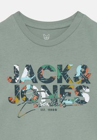 Groene t-shirt met vet, kleurrijke tekst "JACK & JONES Essentials" met spettereffecten en "EST. 1990" eronder.