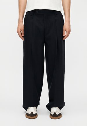 Άνδρας που φορά χαλαρά μαύρα παντελόνια με φαρδιά μπατζάκια και λευκά αθλητικά με καφέ σόλες, στέκεται μπροστά σε μια απλή φόντο.