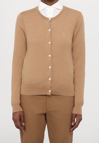 Světle hnědý kardigan vyrobený z měkkého pleteného materiálu, s kulatým výstřihem, zapínáním na knoflíky a malým vyšitým logem na hrudi.