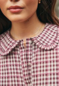 Short-sleeve, buttoned blouse with a frilled collar. Features a burgundy and white checkered pattern. Soft fabric with a textured appearance.