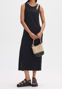 Robe maxi noire sans manches avec un col rond et des découpes sur les côtés, associée à un sac tissé noir et beige et des sandales noires à semelle épaisse.