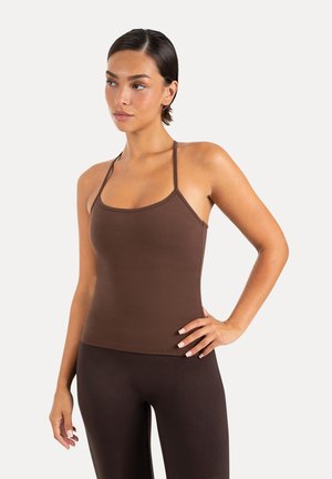 Kvinde med kort mørkt hår iført en tætsiddende brun tanktop og mørkebrune leggings, poserer med den ene hånd på hoften mod en ensfarvet baggrund.