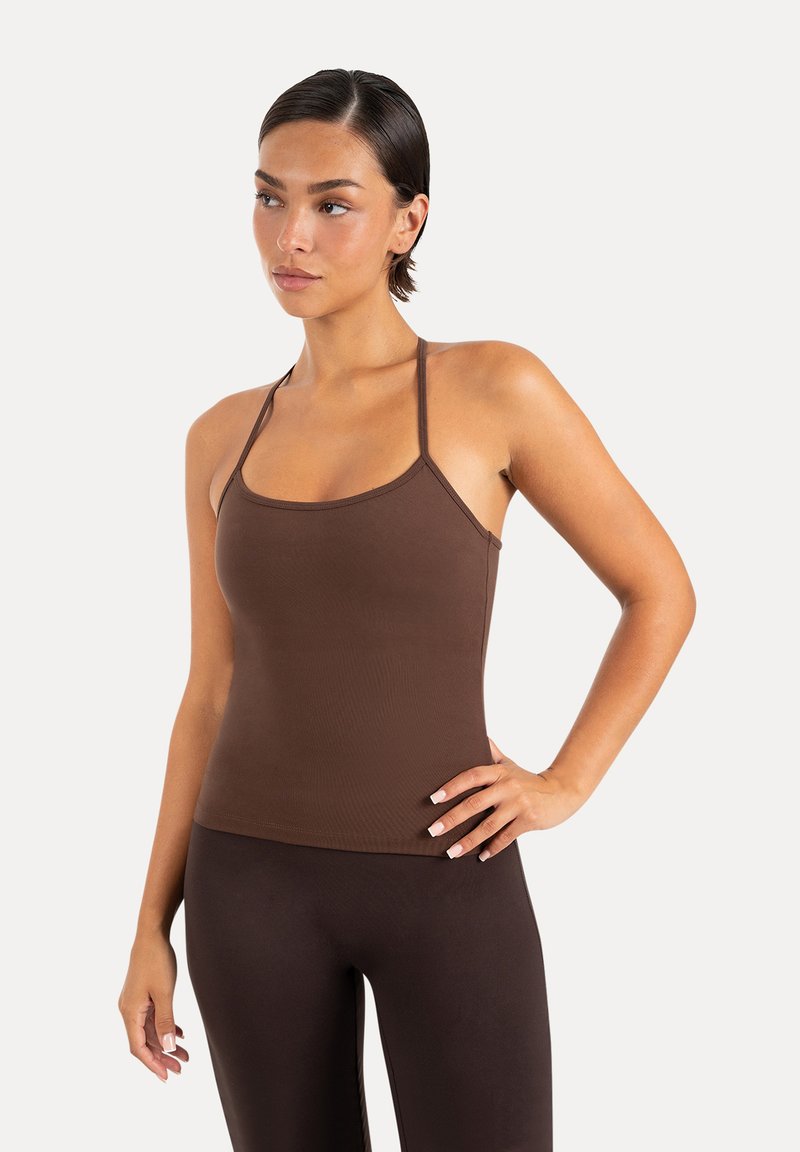 Femme aux cheveux courts et foncés portant un débardeur marron ajusté et des leggings marron foncé, posant avec une main sur la hanche devant un fond uni.