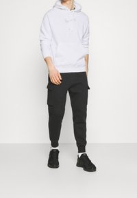 Vit huvtröja med känguruficka och grå logotyp, kombinerad med svarta cargobyxor med sidofickor. Svarta sportskor.