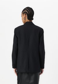 Blazer a righe nere con design su misura, larghe alette e polsini a due bottoni, mostrato dal retro su uno sfondo neutro.