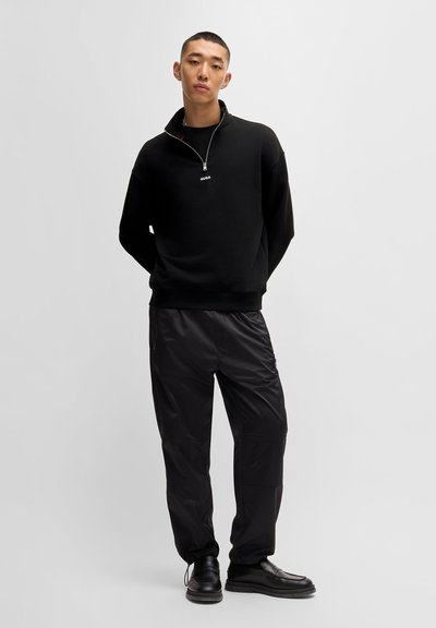 Sweat à fermeture éclair noir avec col montant et logo, associé à un pantalon noir brillant et fuselé, ainsi qu'à des chaussures noires. Fond uni.