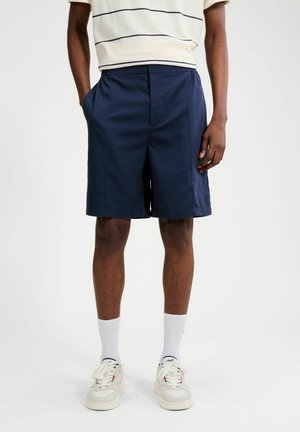 Hombre con pantalones cortos azul marino hasta la rodilla, zapatillas blancas, calcetines blancos hasta la pantorrilla y una camisa blanca de manga corta con finas rayas horizontales.