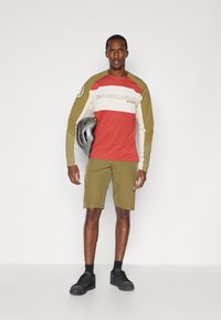 Maglia da ciclismo a maniche lunghe rossa, bianca e verde oliva con la scritta "ENDURA", abbinata a pantaloni corti verdi oliva. Un uomo tiene un casco grigio.