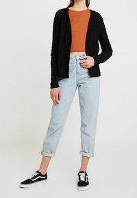 Black knit cardigan layered over an orange ribbed top, paired with light blue cuffed jeans and black sneakers with white accents.