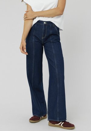 Femme portant un jean large taille haute bleu foncé et des baskets bordeaux, se tenant contre un fond clair uni.