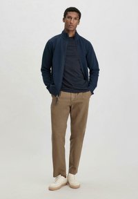 Marinefarbener Reißverschlussjacke über einem dunklen T-Shirt, kombiniert mit khakifarbenen Hosen und weißen Turnschuhen. Glatter Stoff, lockere Passform und lässiges Design.