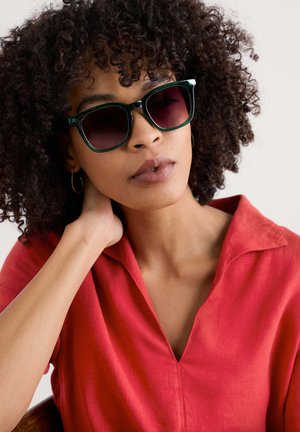 Green cat-eye sunglasses with dark gradient lenses. Worn by a person with curly hair in a red linen shirt, featuring a collar and button detail.
