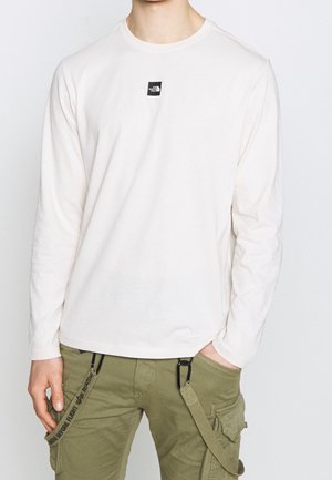 Person trägt ein weißes Langarmshirt mit Rundhalsausschnitt und kleinem schwarzem Logo auf der Brust sowie olivgrüne Cargo-Hosen mit Riemen.