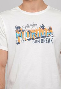 T-shirt en coton blanc avec un graphique coloré portant l'inscription "Salutations du Florida Sun Break" accompagné de designs de palmiers et de fleurs.