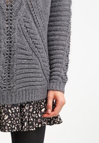 Gray knitted sweater with textured patterns, ribbed cuffs, and a relaxed fit, layered over a black skirt with a white leopard print.