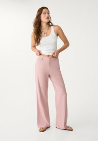 Široké růžové kalhoty vyrobené z lehké tkaniny, zkombinované s bílým topem na ramínka. Modelka doplňuje svůj outfit vícebarevným náhrdelníkem.