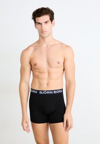Boxer shorts noirs en coton avec une texture lisse, dotés d'une ceinture élastique de marque en bleu marine avec des lettres blanches.