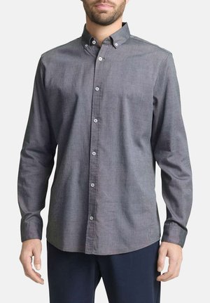 Mann trägt ein graues, langärmeliges Hemd mit Knöpfen am Kragen und dunkle Hosen, steht vor einem einfarbigen Hintergrund.