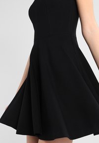 Tailliertes schwarzes ärmelloses Kleid mit weit ausschwingendem Rock, gezeigt in der Seitenansicht von der Taille bis zur Mitte des Oberschenkels auf einer Person mit hellerer Hautfarbe.