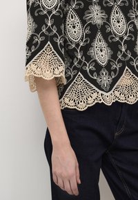 Svart blus med intrikata vita blommor och paisleymönster broderade, med en vågig krämfärgad spetskant. Kombinerad med mörkblå denim.